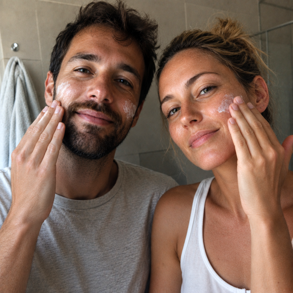 A man and a woman in a bathroom setting, both applying white face cream to their faces.
